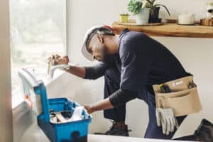 %Title% | 3 Mountains Home Services A 3 Mountains Home Services plumber fixes a bathtub faucet using tools from a blue toolbox in a bright, clean bathroom.