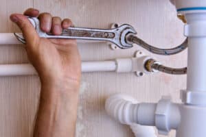 A 3 Mountains Home Services technician tightens a flexible metal pipe with a wrench beneath a customer’s sink, ensuring secure plumbing.