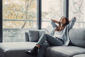 %Title% | 3 Mountains Home Services A woman sits on a couch relaxing with her hands behind her head. Behind the couch is a wall of windows showing a view of leafy green trees.