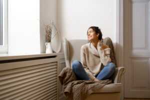 “Smiling woman relaxing indoors in fall, enjoying a home made cozy by a heating system that benefits from professional furnace maintenance.