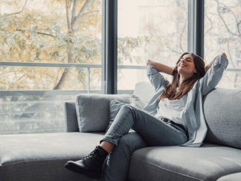 A woman sits on a couch relaxing with her hands behind her head. Behind the couch is a wall of windows showing a view of leafy green trees.